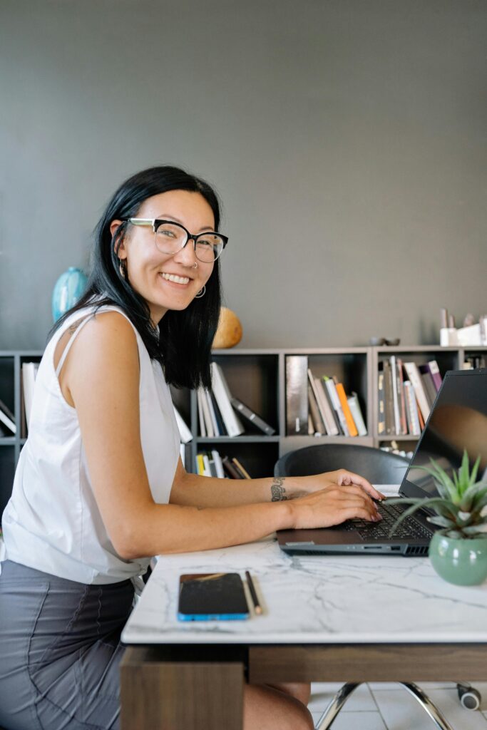 Smiling businesswoman with eyeglasses using laptop at a modern desk in an office environment.